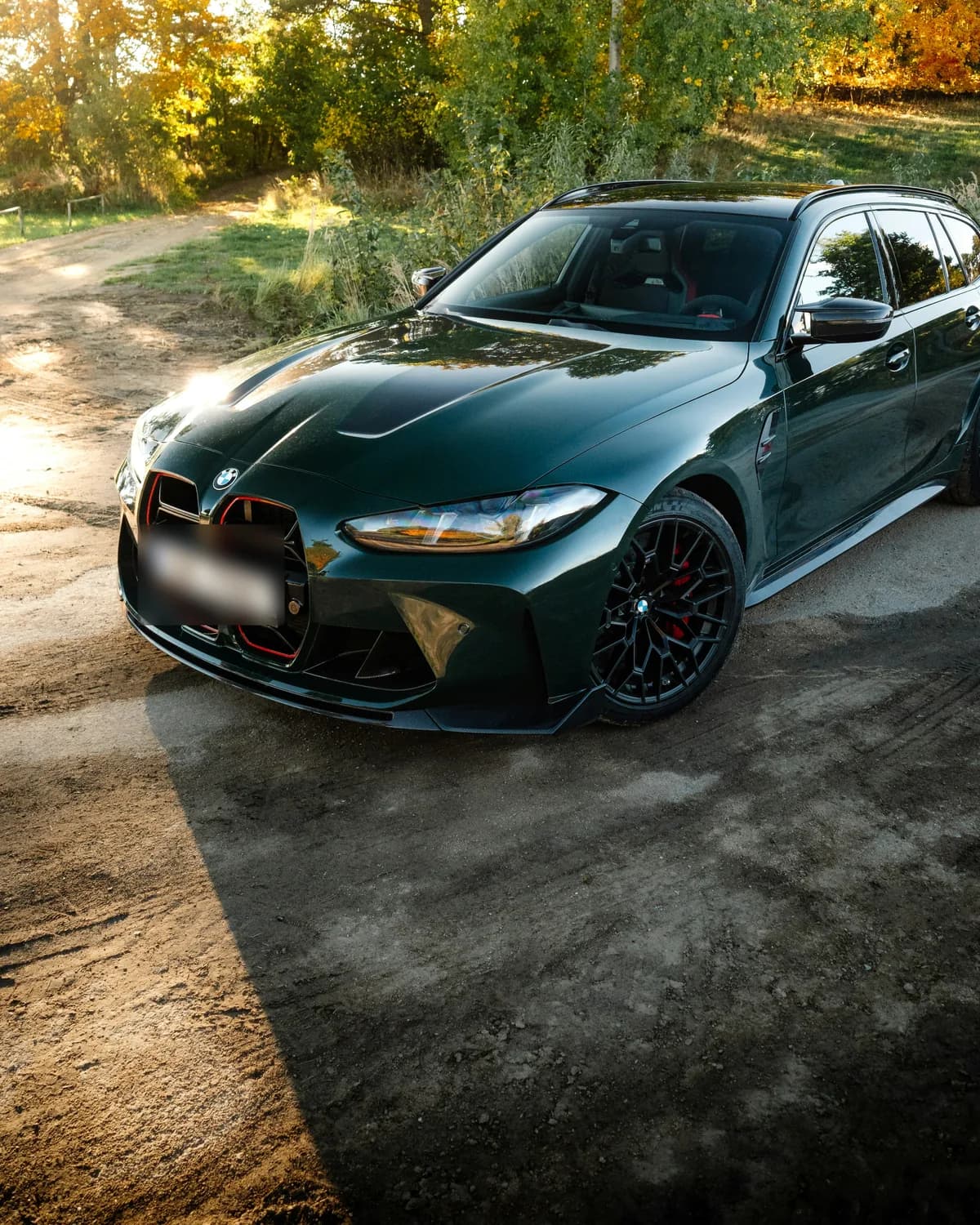 Dark green BMW M3 Touring on a dirt lot in low autumn sun
