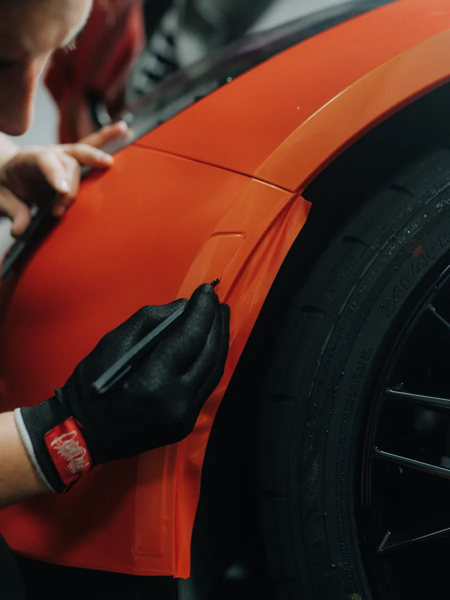 Gloved hand tucking orange vinyl wrap at a wheel arch